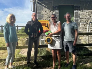 Four adults standing and smiling by a wooden fence in front of a brick building, with one person holding an automated external defibrillator (AED) and a yellow sign for the life saving device displayed in the background.