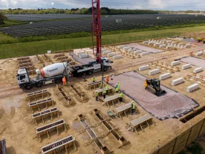 Construction site with workers in high visibility clothing preparing concrete foundations using trucks and machinery. Rectangular molds and blocks are arranged across the dirt ground. In the background, rows of solar panels stretch into the distance beside green farmland.