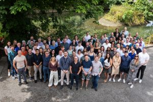 Large group of people posing together outdoors, with trees and a pond in the background.