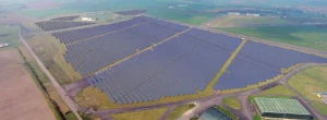 A wide aerial view of an extensive solar farm with multiple large sections of solar panels arranged in rows, covering vast areas of green grass. Surrounding the solar field are access roads, some buildings, and patches of open countryside under a slightly hazy sky.