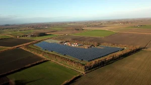 Aerial view of a large solar farm bordered by trees and surrounded by agricultural fields. The solar panels are arranged in long rows, and several small buildings and structures are present near the centre. The landscape is flat with patchwork fields extending into the distance under a clear sky.