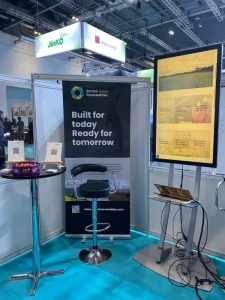 Exhibition booth for British Solar Renewables featuring a banner, tall table with snacks, stool, and a digital display, with overhead signs from other companies.