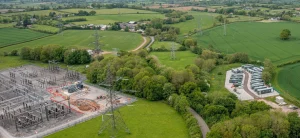 Aerial view of a rural landscape featuring a large electrical substation with metal pylons on the left, and rows of battery storage containers on a paved site to the right. Both facilities are surrounded by green fields, trees, and hedges, with country roads and scattered farm buildings visible in the background.