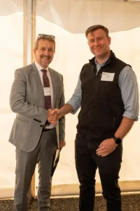 Two people stand in front of a light coloured tent or marquee, shaking hands. One person wears a light grey suit, patterned tie, and glasses, while the other wears a black vest over a blue shirt with rolled sleeves and a black watch.