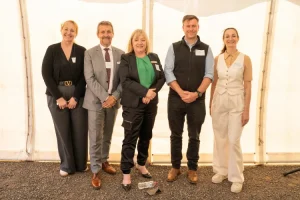 Five people stand side by side in front of a white tent or marquee. They are dressed in business and smart casual attire, wearing name badges.