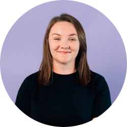 Portrait of a woman with straight light brown hair, wearing a plain black t-shirt, seated in front of a light purple background.